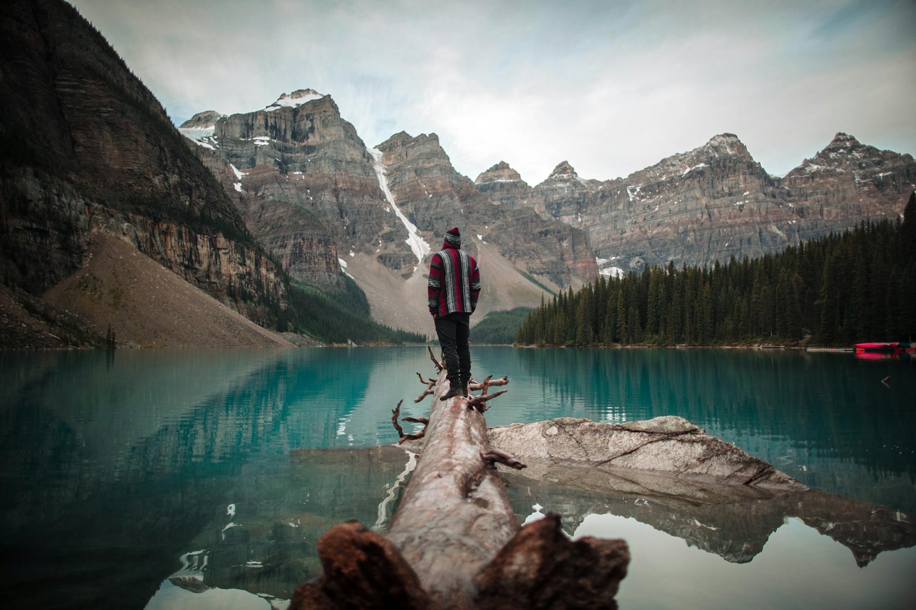 man wearing red and white jacket standing on brown logs looking at lake and icy mountain scenery